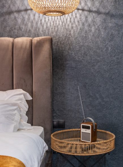 A bedside table holding a small radio next to a bed with a brown suede headboard.